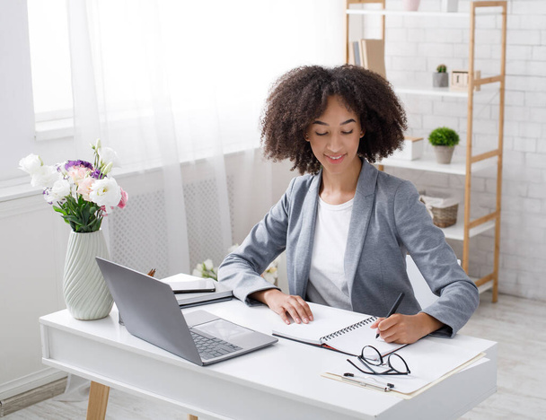 Adopt an order from clients in online office at home during outbreak. Cheerful african american female manager works at table with laptop and writes in notebook - 写真・画像