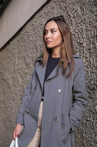 attractive young asian model in gray trench coat standing near wall outdoors - Photo, Image