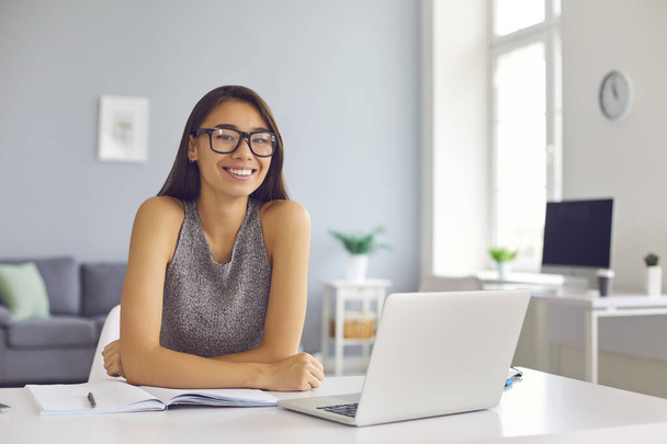Happy young woman in cool eyeglasses sitting at desk with open laptop and notebook looking at camera - Foto, imagen