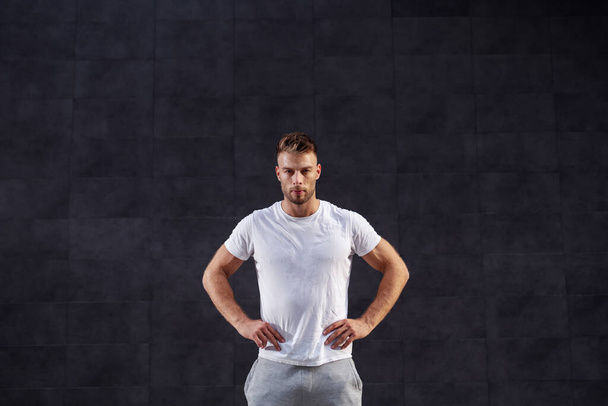 Pohledný svalnatý blonďák ve sportovním oblečení pózuje venku s rukama v bok. V pozadí je šedá zeď. - Fotografie, Obrázek