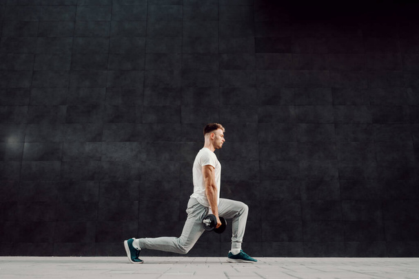 Full length of handsome fit muscular caucasian blond bearded man in tracksuit and t-shirt doing lunges with dumbbells in hands. - Photo, Image