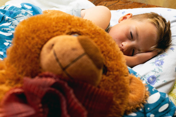 Photo of little cute schoolboy sleeping with big brown teddy ber in bed. Eyes are closed. - Fotoğraf, Görsel