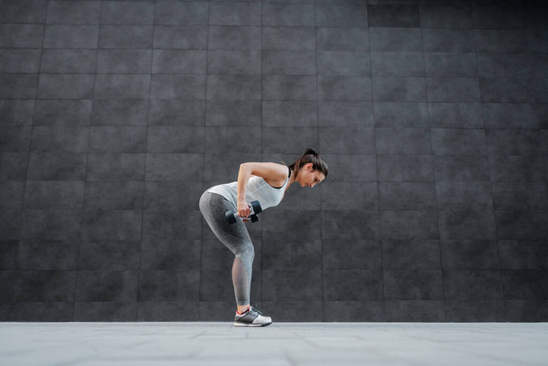 Side view of fit healthy caucasian brunette in sportswear bending and doing exercises with dumbbells in hands. In background is dark wall. - Valokuva, kuva