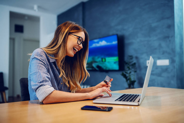 Side view of young charming caucasian woman with eyeglasses holding credit card and using laptop for online shopping while sitting at dining table at home. - Foto, Imagem