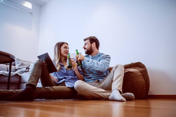 Mladý atraktivní bělošský pár sedící v obývacím pokoji na podlaze a připíjející si pivem. Žena držící tableta. - Fotografie, Obrázek