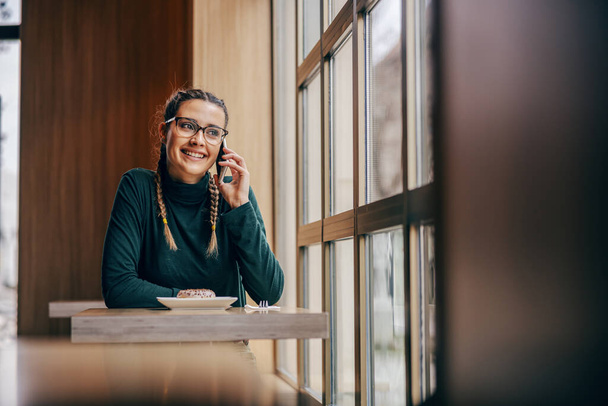 Sorridente giovane donna seduta in mensa accanto alla finestra e avendo conversazione telefonica. Concetto di nuove tecnologie. - Foto, immagini