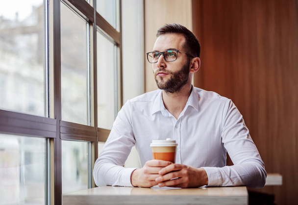 Young thoughtful man elegantly dressed sitting in fast food restaurant, holding disposable cup of coffee and looking trough window. He is having lunch break. - Foto, Imagem