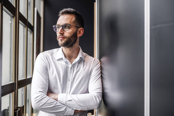 Young serious attractive elegant bearded businessman standing near window with arms crossed and looking trough it. - Photo, Image