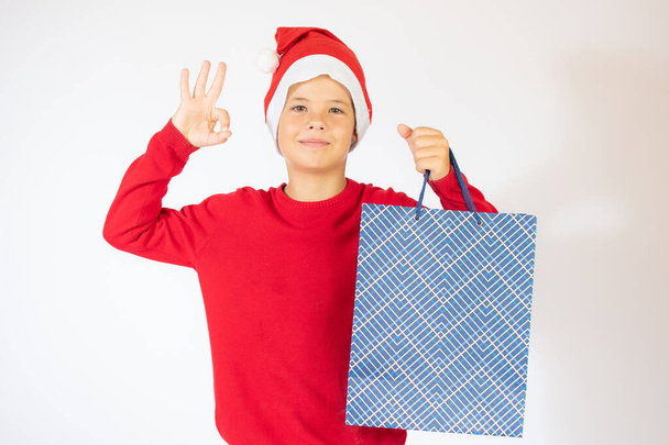 Portret uśmiechniętego chłopca w kapeluszu Świętego Mikołaja, zaskoczony. Biały w czerwonym swetrze, z torbami na zakupy. Święta koncepcja Bożego Narodzenia - Zdjęcie, obraz