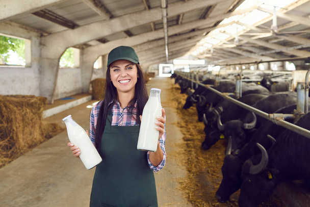 Happy young woman standing in cowshed, holding bottles of fresh milk and looking at camera - 写真・画像