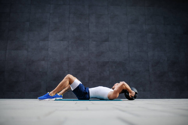 Side view of handsome muscular caucasian man is shorts and t-shirt lying down on mat and doing exercises with kettle bell. - Fotografie, Obrázek