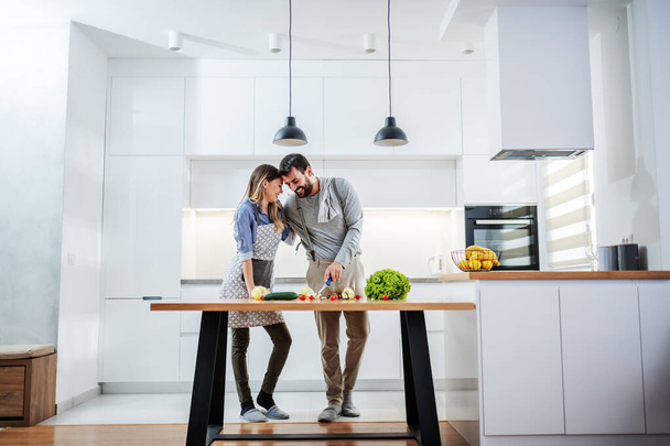 Young attractive caucasian bearded man hugging his beloved girlfriend and cutting vegetables for meal. Healthy eating concept. - Photo, Image