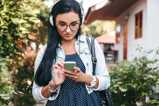 young beautiful stylish woman using smartphone, headphones, eye glasses, summer, vintage denim outfit, smiling, happy, positive - Foto, Imagen