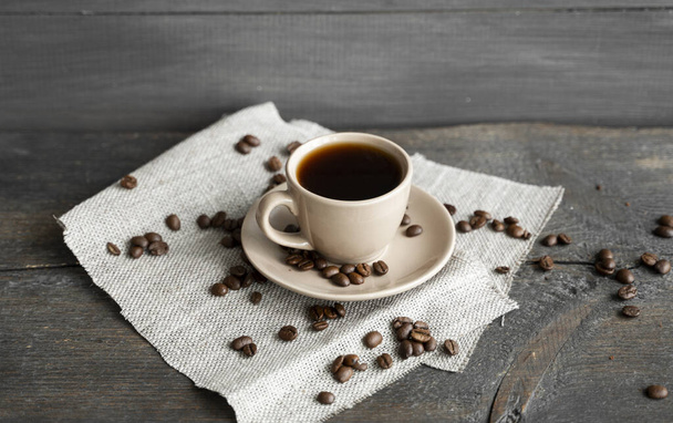 Coffee cup with scattered coffee beans on a wooden table background. Mug of black coffee on a linen textile. - Photo, image