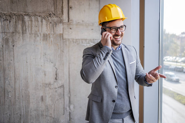 Lächelnder Architekt im Anzug und mit Helm auf dem Kopf steht im Bauprozess neben dem Fenster und telefoniert mit dem Smartphone. - Foto, Bild