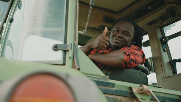 Young african farmer sitting in the tractor cabine and showing thumbs up as approval - Foto, afbeelding