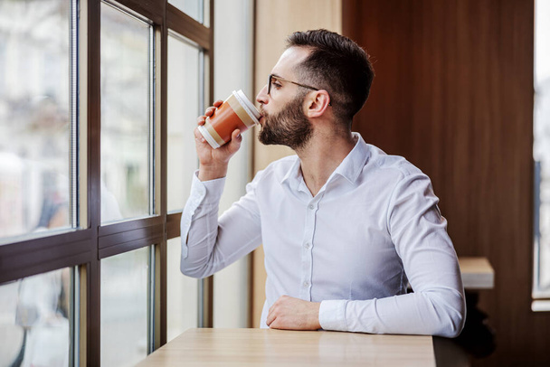 młody geeky człowiek elegancko ubrany siedzi w kawiarni obok okna i pije kawę z jednorazowego kubka. - Zdjęcie, obraz