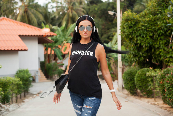 young stylish hipster woman in black t-shirt, jeans, listening to music on headphones, having fun, walking in the street, summer vacation, enjoying - Foto, Bild