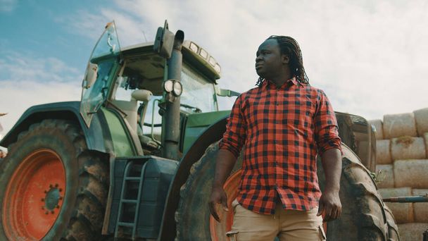 Afrikalı bir adam, yeşil traktörün önünde duran bir çiftçi. Arka planda saman yığınları - Fotoğraf, Görsel