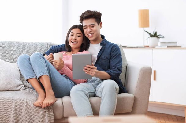 Para azjatycka za pomocą tabletu cyfrowego siedzi na kanapie w domu - Zdjęcie, obraz