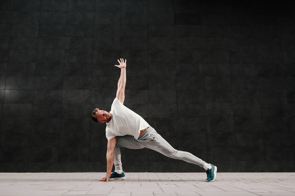 Strong muscular handsome caucasian bearded blonde man in tracksuit doing stretching exercises for backs. In background ins gray wall. - Photo, Image