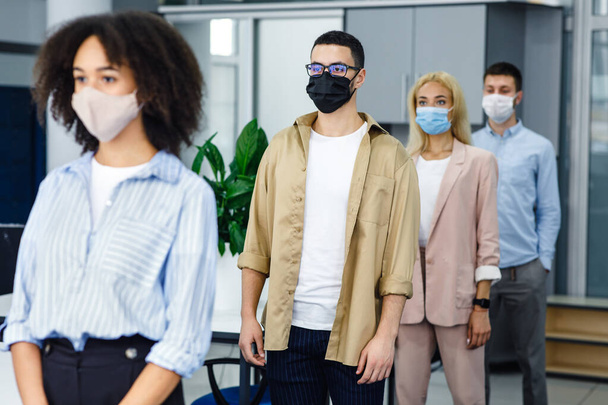 仕事と新しい通常の前に温度をチェックするキュー。コロナウイルス流行時に職場の入り口付近の保護マスクの多人種労働者 - 写真・画像