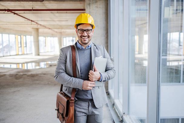 Молодой улыбающийся архитектор, стоящий у окна в здании в процессе строительства со шлемом на голове и ноутбуком под мышкой и смотрящий в камеру. - Фото, изображение