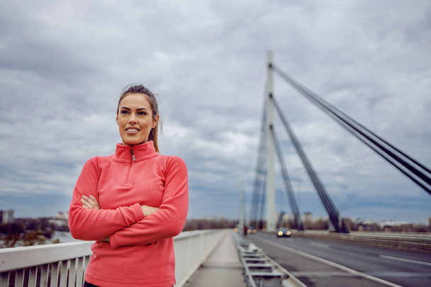 Ylpeä kaunis hymyilevä urheilija urheiluvaatteissa seisoo sillalla kädet ristissä ja katselee kameraa. Fitness pilvinen sää käsite. Kaupunkien elämäntapa. - Valokuva, kuva