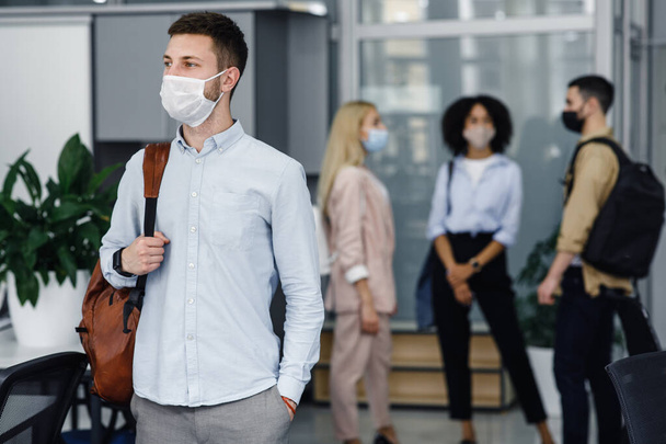 Social distance and work in office after covid-19 quarantine. Focus on millennial man with protective mask and backpack, looks to the side in hallway - Photo, Image