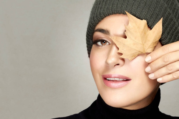 Autumn concept. Beautiful young woman in warm polo neck top and beanie holding a dried maple leaf to her eye smiling at camera. Beauty portrait over a grey background with copyspace - Photo, Image