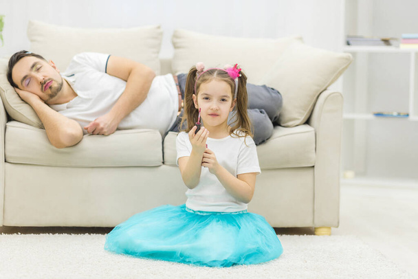 Photo of father sleeping on sofa and his daughter sits on white floor. - Foto, afbeelding