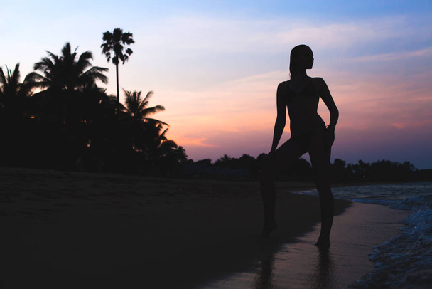 young beautiful slim woman standing on beach at dawn, tropical vacation, palm tress, silhouette, sexy, sensual, ocean waves, colorful sky - Фото, зображення