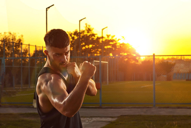 Athlete trains on a sunset background. The boxer warms up near the stadium. Contrasting dramatic shadows on the face as an artistic effect. The guy on the playground. - Fotografie, Obrázek