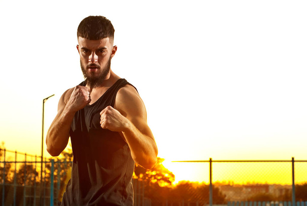 Athlete trains on a sunset background. The boxer warms up near the stadium. Contrasting dramatic shadows on the face as an artistic effect. The guy on the playground. - Φωτογραφία, εικόνα