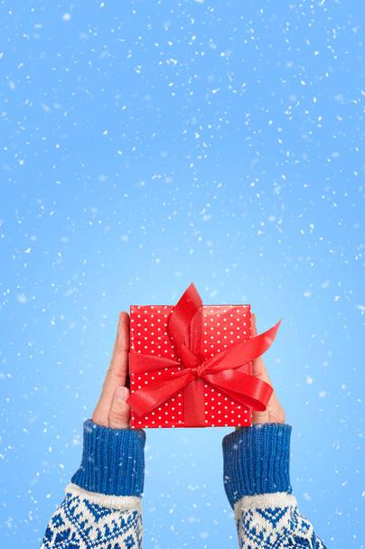 Hands is holding a red gift box with ribbon at blue background with snow. Merry Christmas card.  - Foto, Imagem