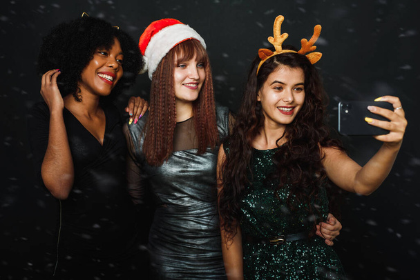 Un gruppo di amiche di diverse nazionalità si fa un selfie natalizio in studio. Giovani donne in cappelli di Babbo Natale e corna di cervo si stanno divertendo insieme - Foto, immagini