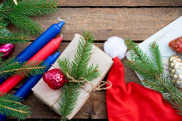 クリスマスツリーの装飾やモミの木の枝、赤いキャップ、キャンドルのクリスマス構成。スプルースの小枝からクラフト紙に包まれた贈り物。濃い木の質感の背景。フラットレイ. - 写真・画像