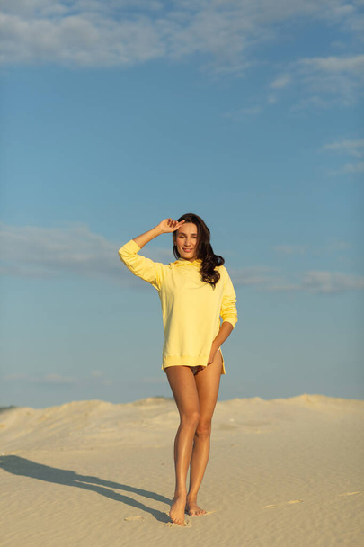 A young beautiful girl with a good figure, around only white sand and blue sky, she is wearing a yellow sweater - Zdjęcie, obraz