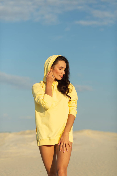 A young beautiful girl with a good figure, around only white sand and blue sky, she is wearing a yellow sweater - Фото, изображение