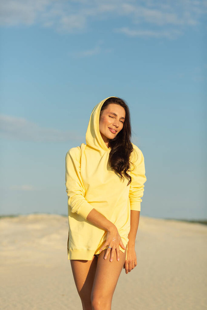 Une jeune belle fille avec une bonne silhouette, autour seulement du sable blanc et du ciel bleu, elle porte un pull jaune - Photo, image