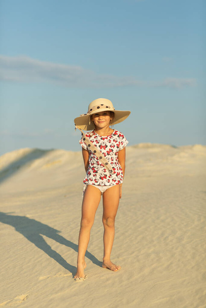 A little girl in the sand, wearing her mother's big hat. all around white sand and blue sky - Foto, Imagem