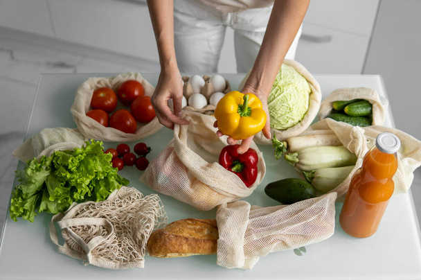 Womans hand, holding a reusable grocery bag with vegetables on a kitchen at home and takes pepper out. Zero waste and plastic free concept. Mesh cotton shopper with vegetables. Ecology. - Foto, immagini