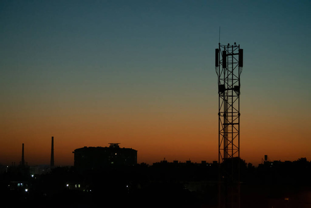 Éjszakai táj, körvonalai városi épületek és kommunikációs torony mobil üzemeltető antenna hátterében naplemente és kék ég - Fotó, kép