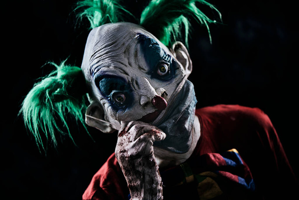 primo piano di un clown cattivo inquietante, con i capelli verdi, con un costume rosso sporco, un papillon sporco e guanti sporchi e sanguinosi, tirando giù o su la sua brutta maschera per il viso, su uno sfondo nero - Foto, immagini