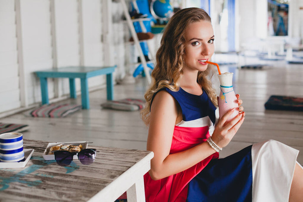 young stylish beautiful woman in sea cafe, drinking cocktail smoothie, sunglasses, flirty, resort style, fashionable outfit, smiling, marine colors dress - Foto, afbeelding