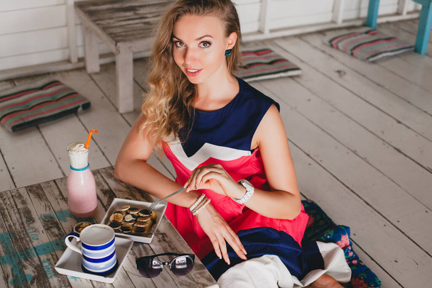 young stylish beautiful woman in sea cafe, eating pancakes, cocktail smoothie, sunglasses, flirty, resort style, fashionable outfit, smiling, marine colors dress - Foto, afbeelding