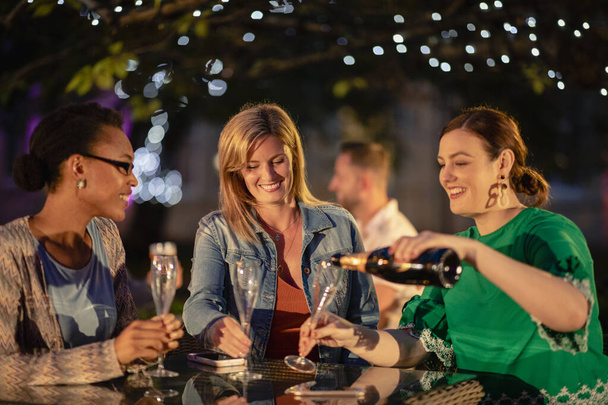 Group of diverse female friends drinking champagne outdoors at a bar/restaurant, celebrating and having fun together. - 写真・画像