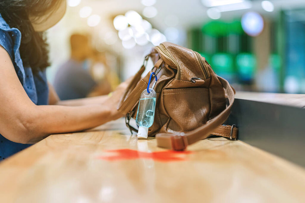Μίνι μπουκάλι αλκοόλ τζελ για να σκοτώσει Corona Virus (Covid-19) κρέμονται σε μια δερμάτινη τσάντα ώμου μιας γυναίκας που φοράει μάσκα, ενώ χρησιμοποιούν smartphone με εναλλακτική σήμανση καθισμάτων για κοινωνική απόσταση στο εμπορικό κέντρο. - Φωτογραφία, εικόνα