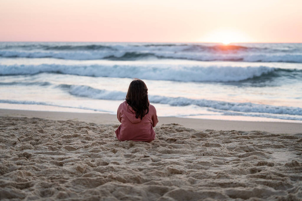 portrait girl in pink sweatshirt looking at the sea sitting on the sand with a lovely background at sunset on the beach - Photo, Image