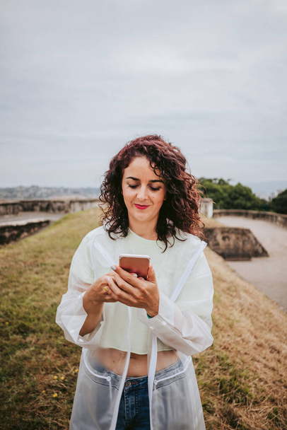 Красивая молодая женщина в белом дождевом пиджаке в интернете на телефоне в дождливый день на севере Испании. Технологии и стиль жизни. - Фото, изображение